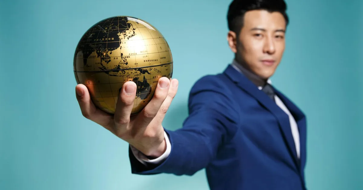Asian businessman in blue suit confidently holding a golden globe against a blue background.