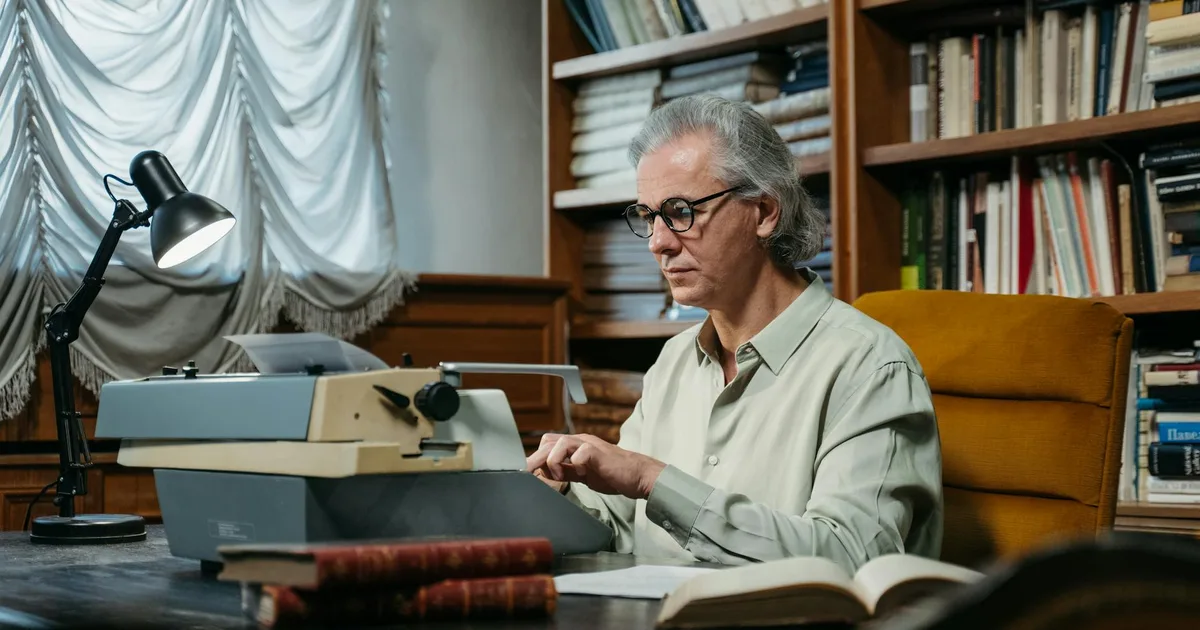 Senior man with glasses and gray hair typing on vintage typewriter in cozy library setting.